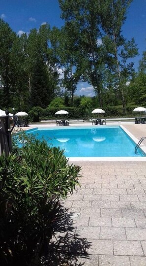 A rectangular swimming pool with a tiled deck, surrounded by lush greenery and trees. There are several white umbrellas and lounge chairs around the pool. - Camping Rio Verde (Costacciaro)