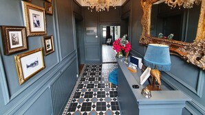 Un pasillo con suelo a cuadros blanco y negro, un candelabro, cuadros en las paredes, un espejo y una recepción con una lámpara azul.
