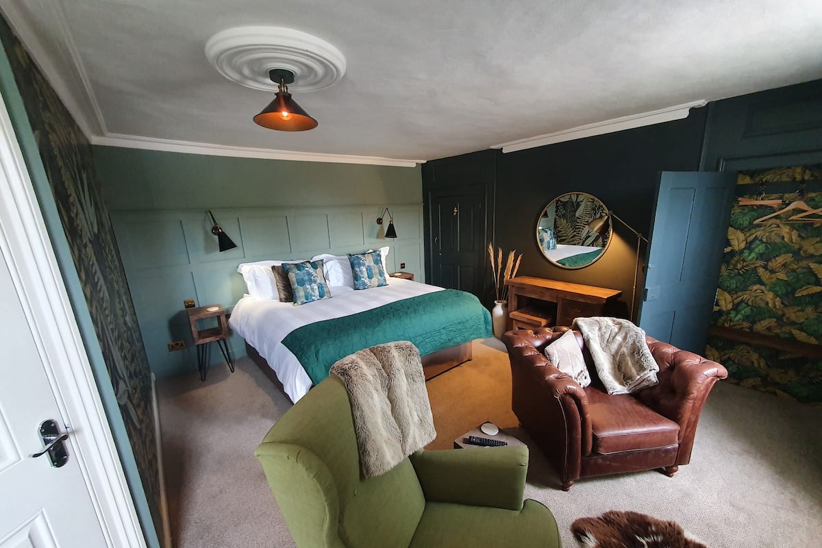Un dormitorio con una cama, un sillón verde, un sillón de cuero marrón, un ventilador de techo y un espejo montado en la pared.