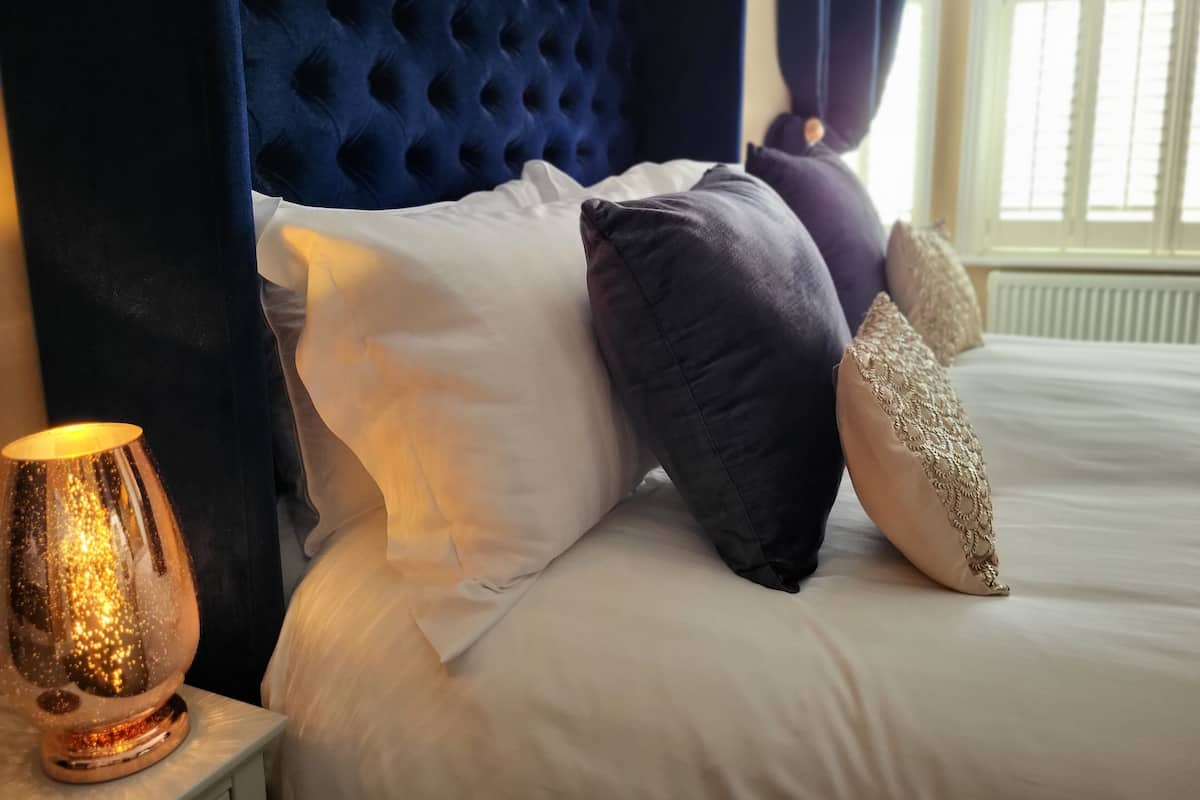 Una cama impecablemente tendida con un cabecero capitonado, almohadas blancas y oscuras, y una lámpara decorativa en una mesita de noche.