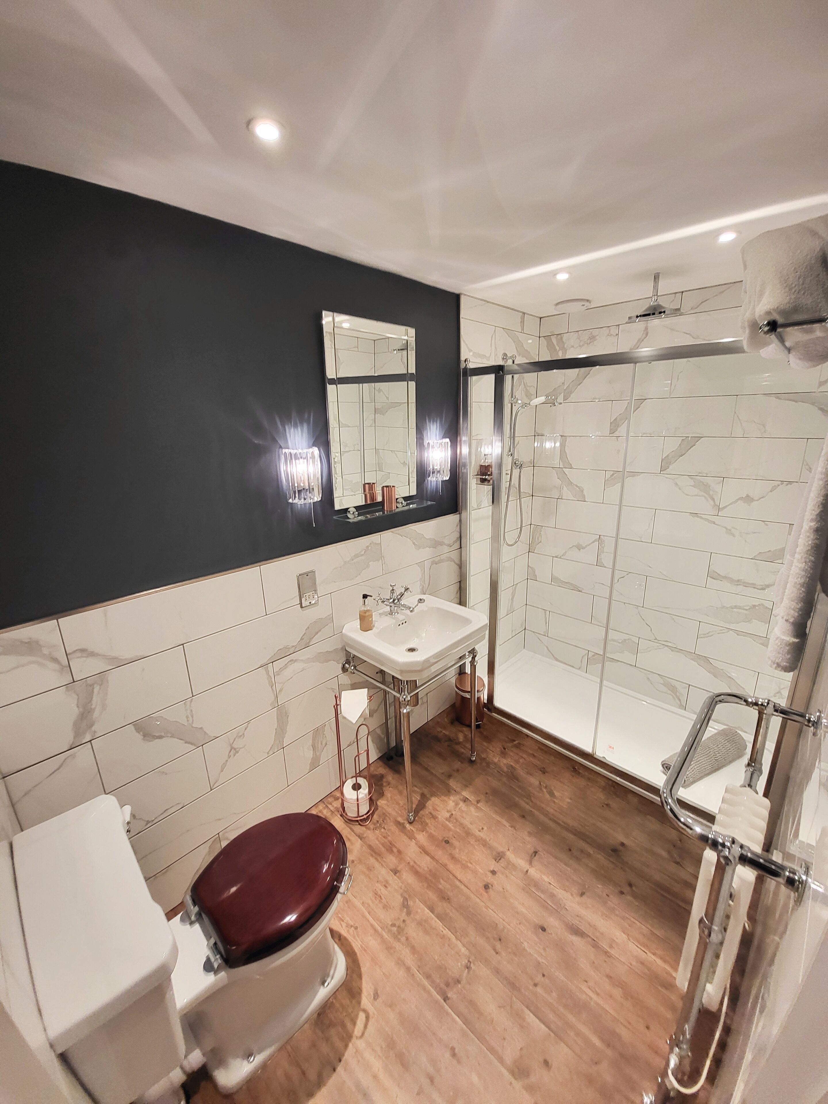 A modern bathroom with a marble-tiled shower, a white sink, and a toilet with a red seat.