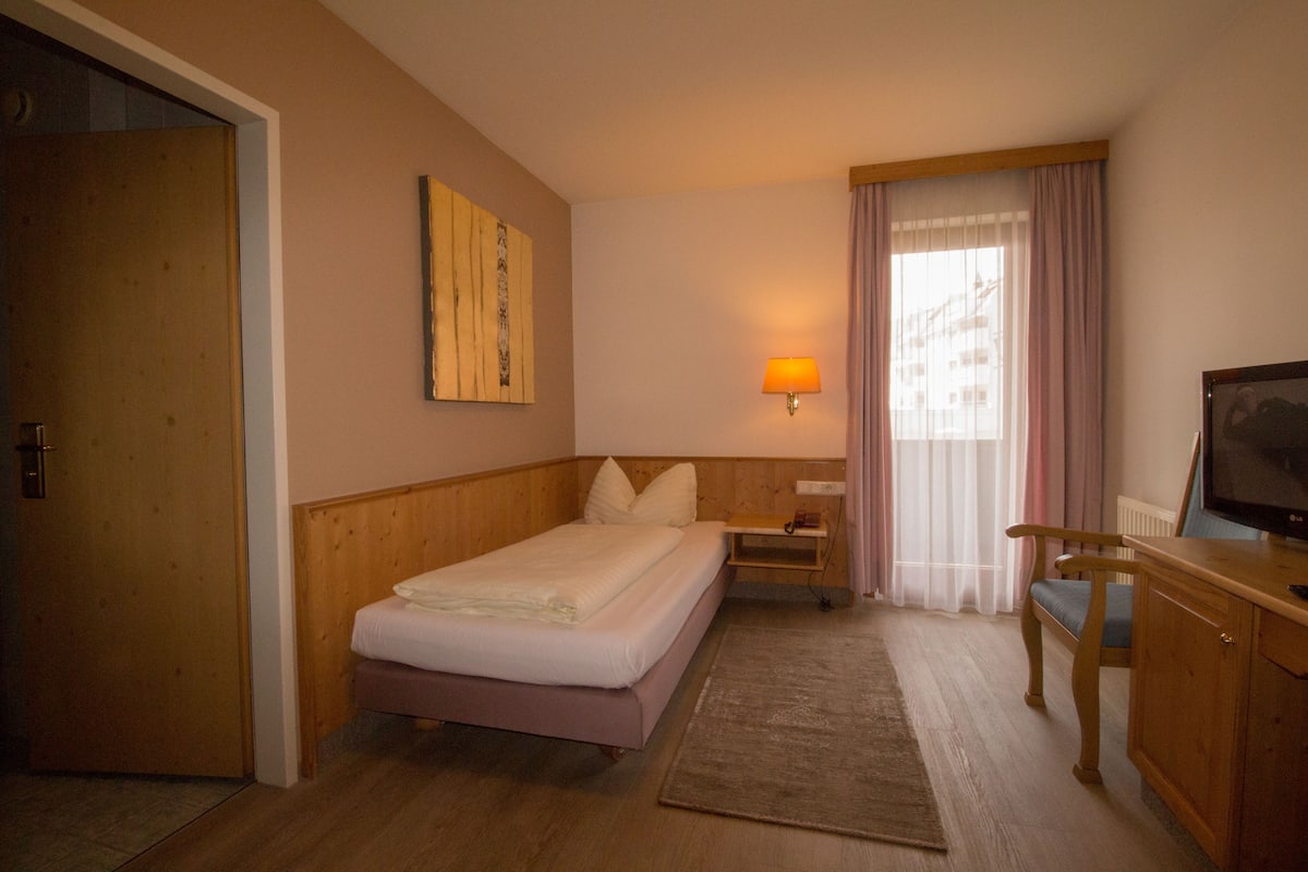 A hotel room with a bed, a desk with a chair, a television, a lamp, and a window with curtains.