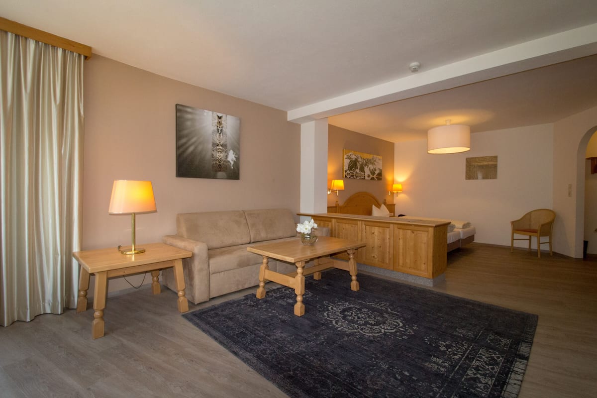 A hotel room with a sofa, a wooden coffee table, a small table with a lamp, a bed, a chair, and a kitchen area.