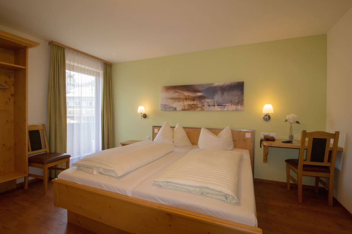 A double bed with two pillows, a bedside table, a chair, a desk, and a painting on the wall.