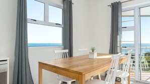Spiseplads med et træbord og hvide stole, store vinduer med grå gardiner og havudsigt.