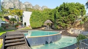 En swimmingpool med træterrasse, omgivet af grøn bevoksning og et bjerg i baggrunden.