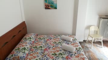 Una cama bien hecha con una colcha floral, un cuadro pequeño en la pared y una silla cerca del radiador.