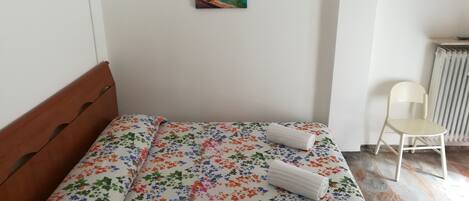 A neatly made bed with a floral bedspread, a small painting on the wall, and a chair by the radiator.