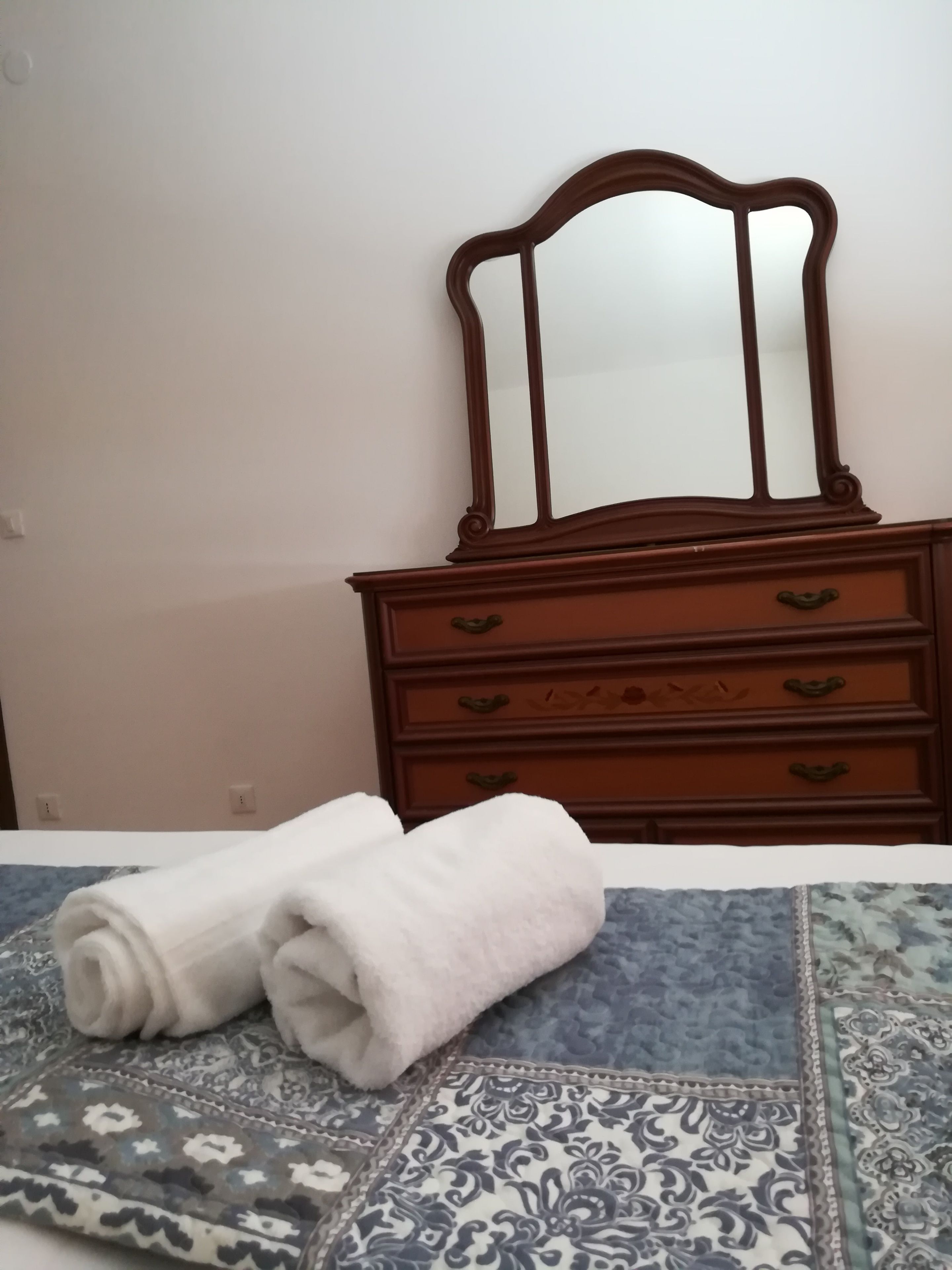 A bed with a patterned bedspread, a wooden dresser with a mirror, and two rolled towels.