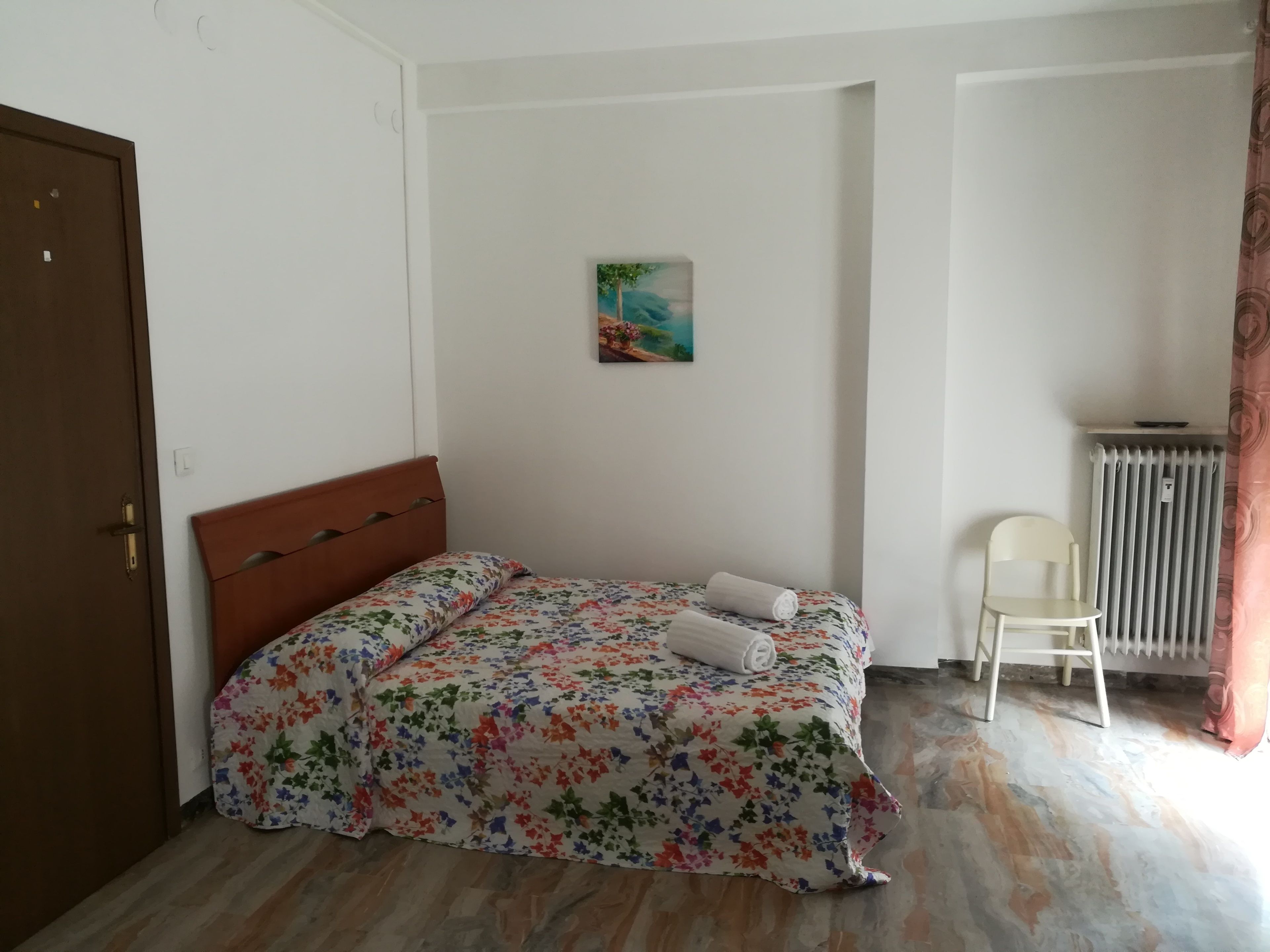 A small, neatly arranged bedroom with a single bed, a chair, a radiator, and a painting on the wall.
