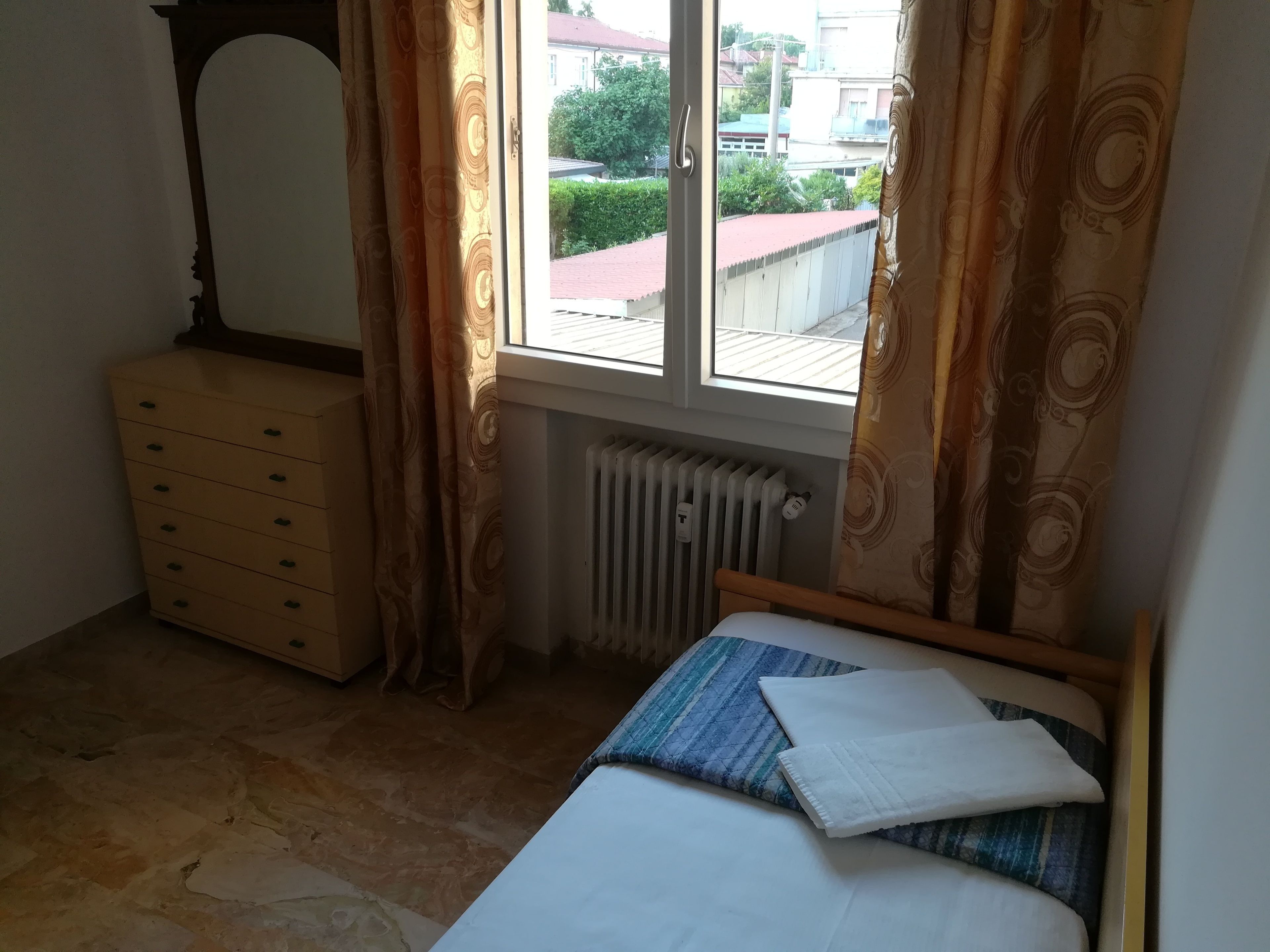 A bedroom with a bed, a dresser, a window with curtains, and a radiator.