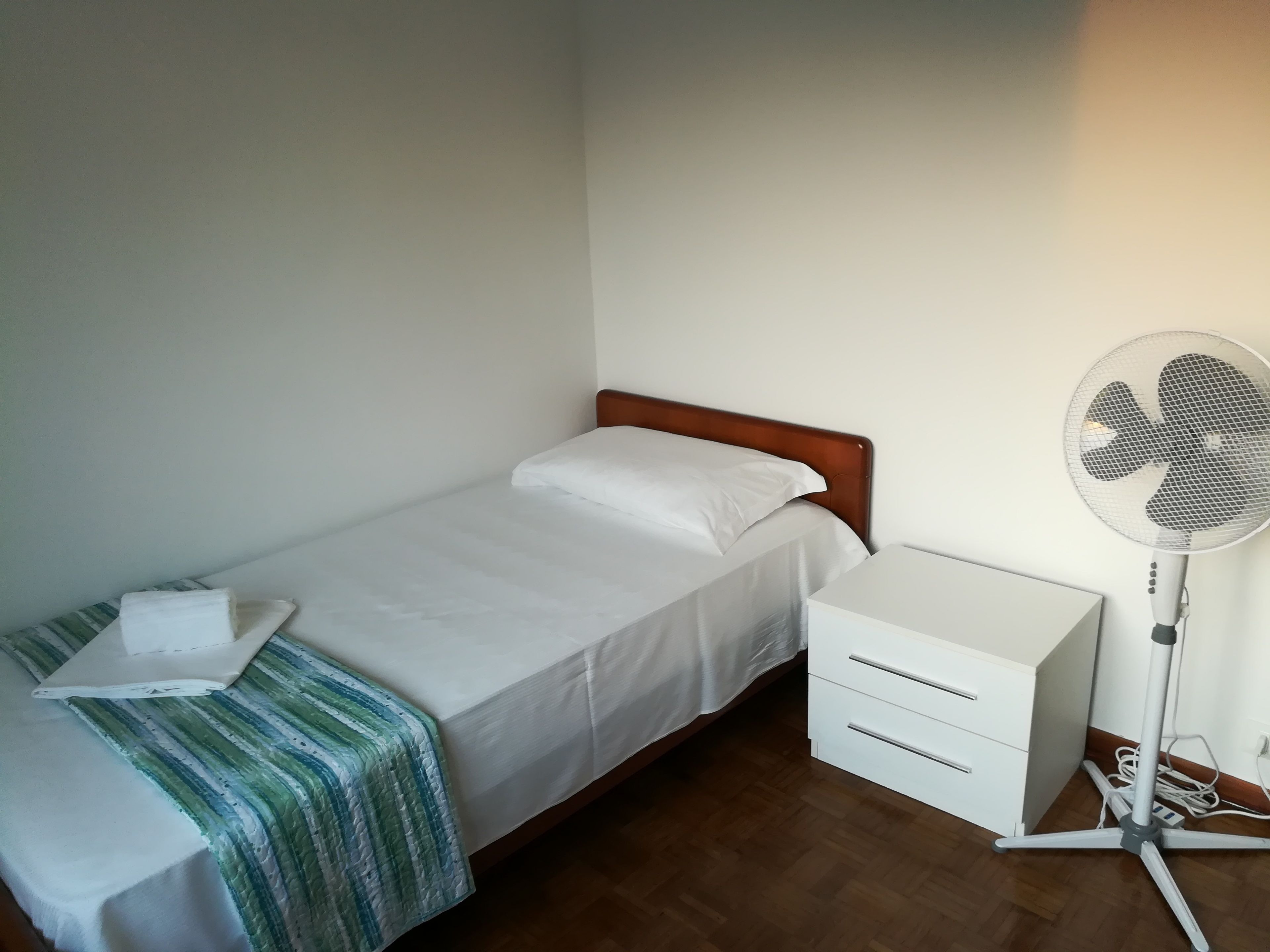 A single bed with a wooden headboard, a bedside table with drawers, and a standing fan.