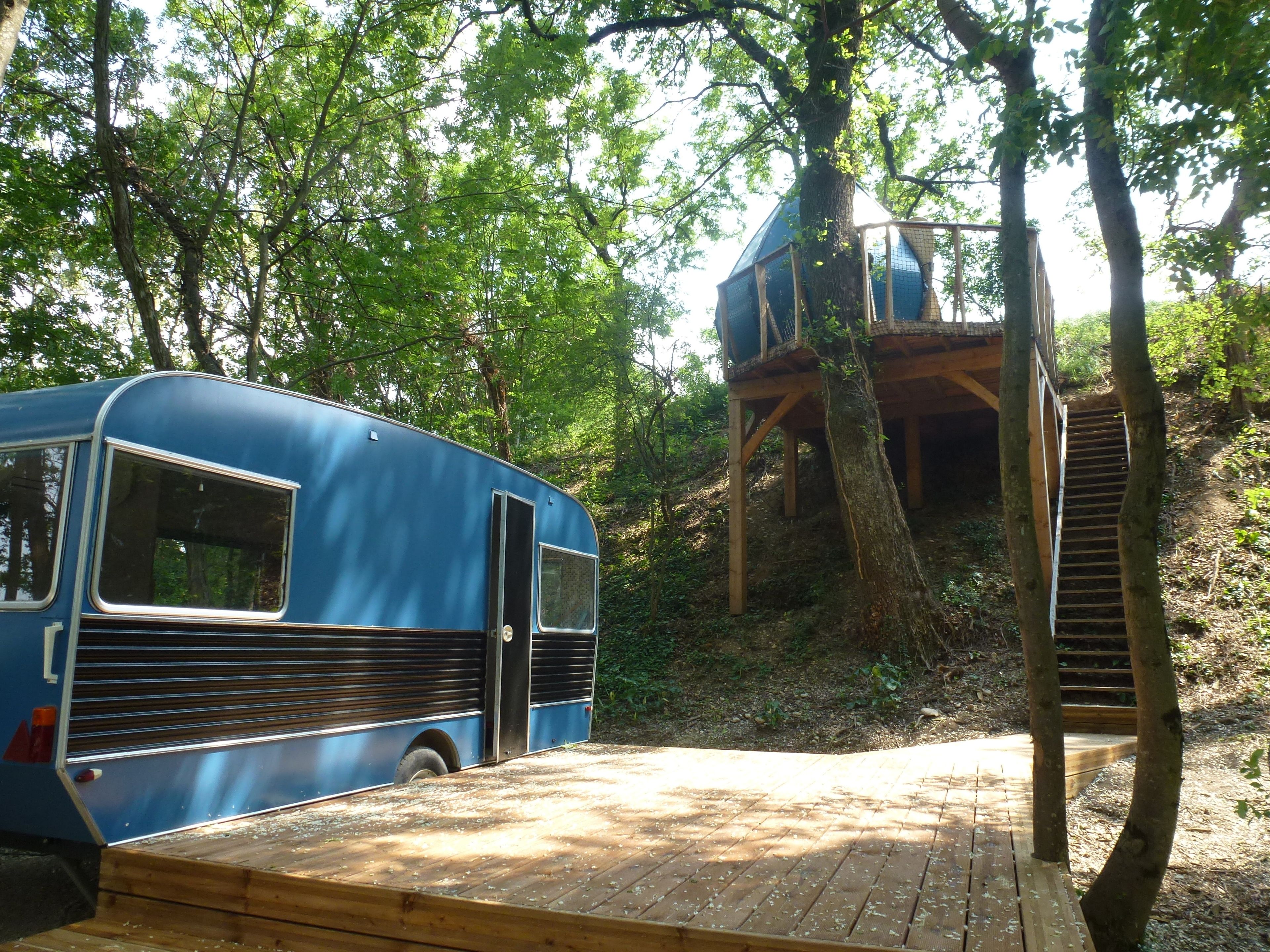 Um ônibus azul estacionado em área arborizada com um deck de madeira e uma casa na árvore.