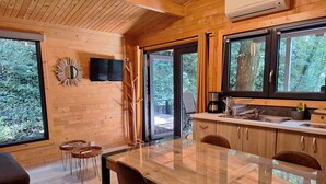 A modern kitchen with wooden cabinetry, a glass dining table, and large windows offering a view of the outdoors. - Des Branches & Vous (Chateauneuf-sur-Isere)