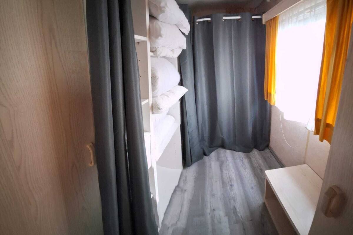 A narrow corridor with a bed on the left, a window with curtains on the right, and a door at the end.
