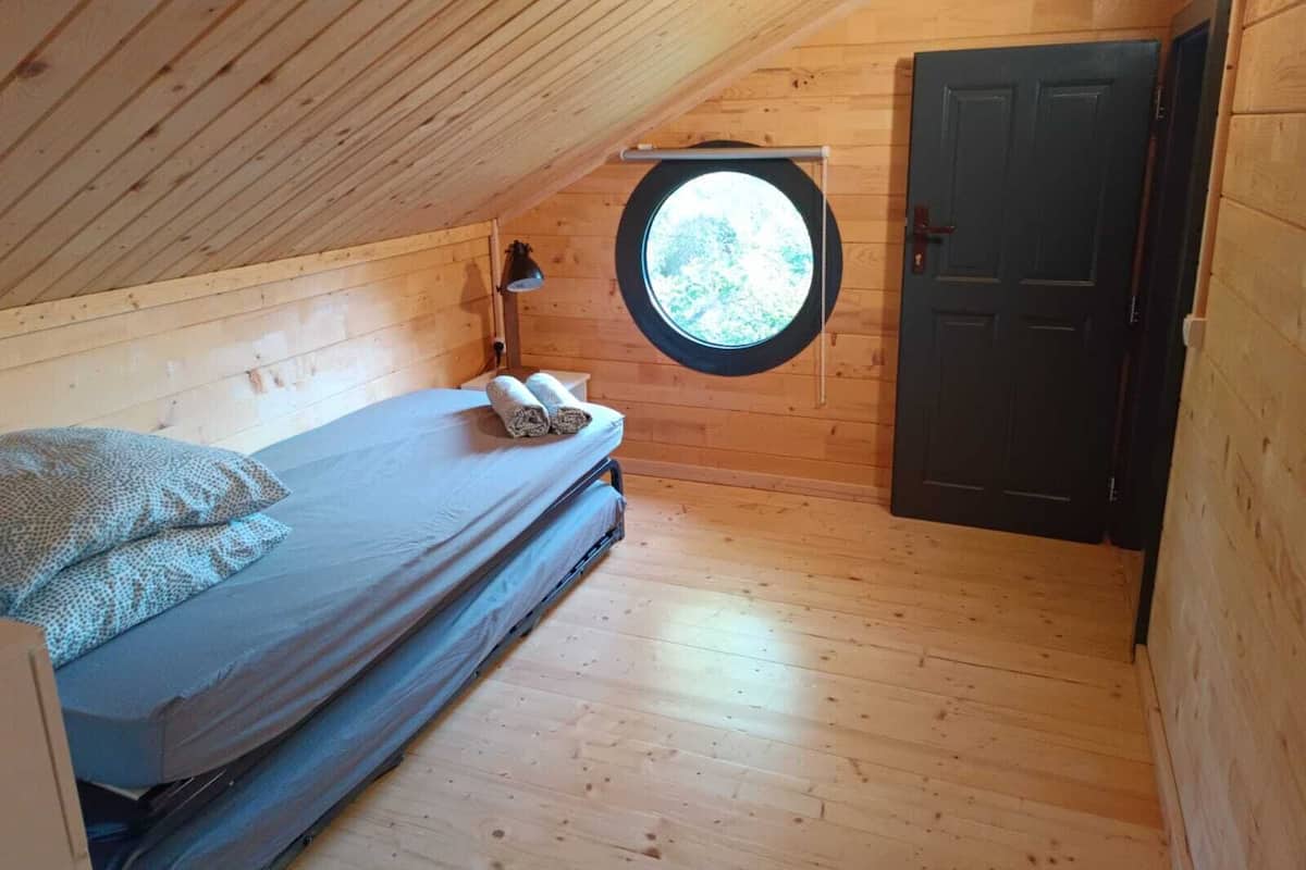A small, wooden room with a single bed, a round window, and a door.