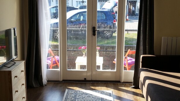 A room with a large sliding glass door, a wooden floor, a dark-colored sofa, a small wooden dresser, and a television on a stand. - 1 Sterling Court City Center (Canterbury)