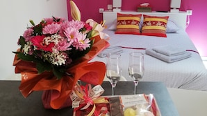 Habitación de hotel con cama, una mesa con un ramo de flores, dos vasos y una caja de regalo.