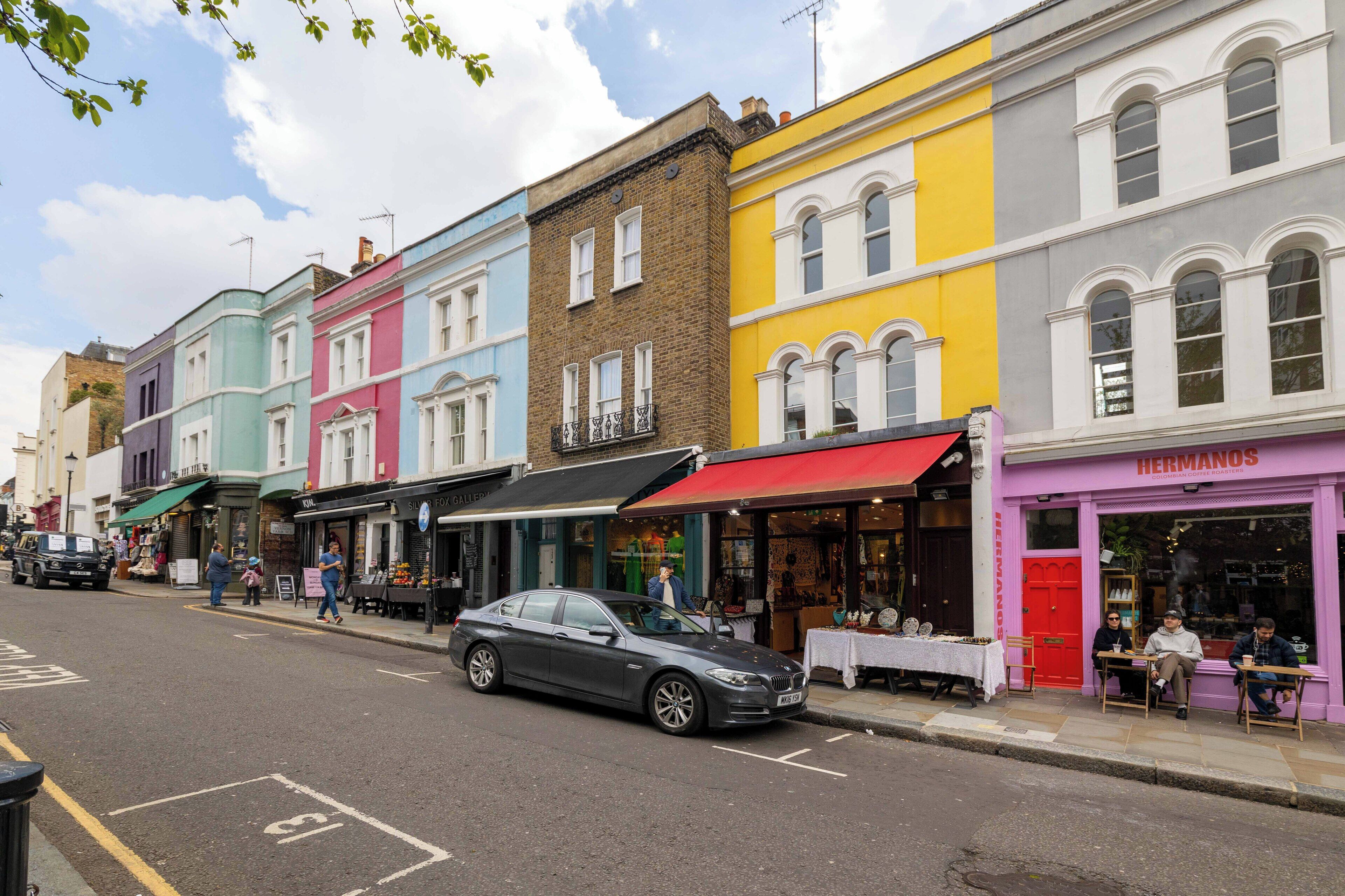 A street with colorful buildings, a parked car, and people sitting outside a shop.