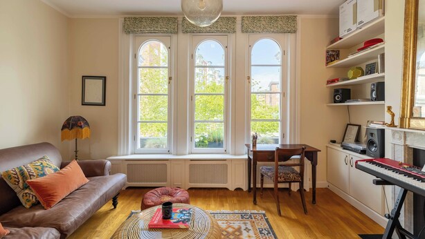 Una sala de estar con un sofá de cuero marrón, una mesa redonda de centro con un libro, un tapete estampado y un asiento junto a la ventana con vistas a edificios y árboles.