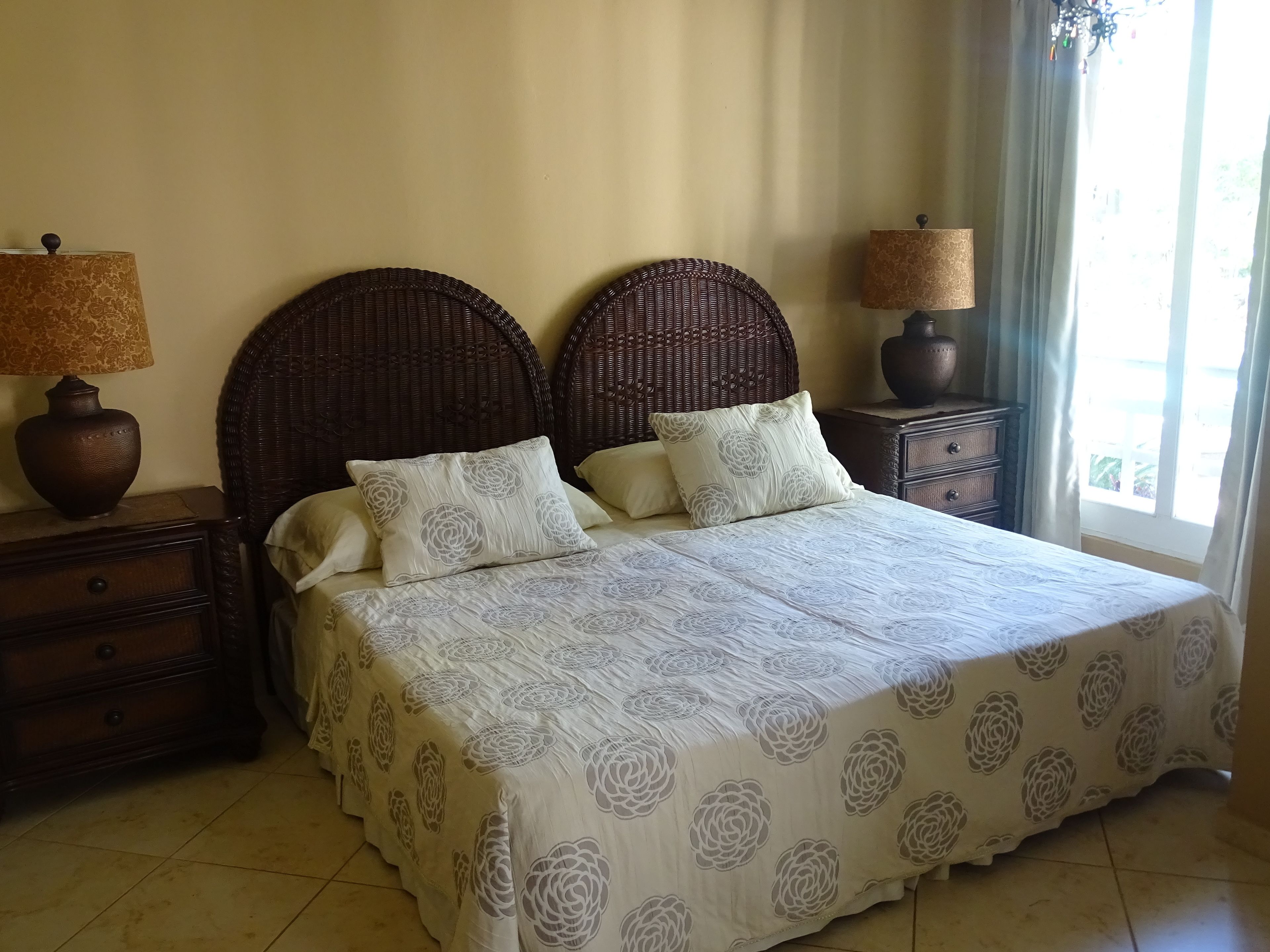 Un lit bien fait, recouvert d’une couvre-lit à motifs floraux, deux tables de chevet avec des lampes et une grande fenêtre avec des rideaux.
