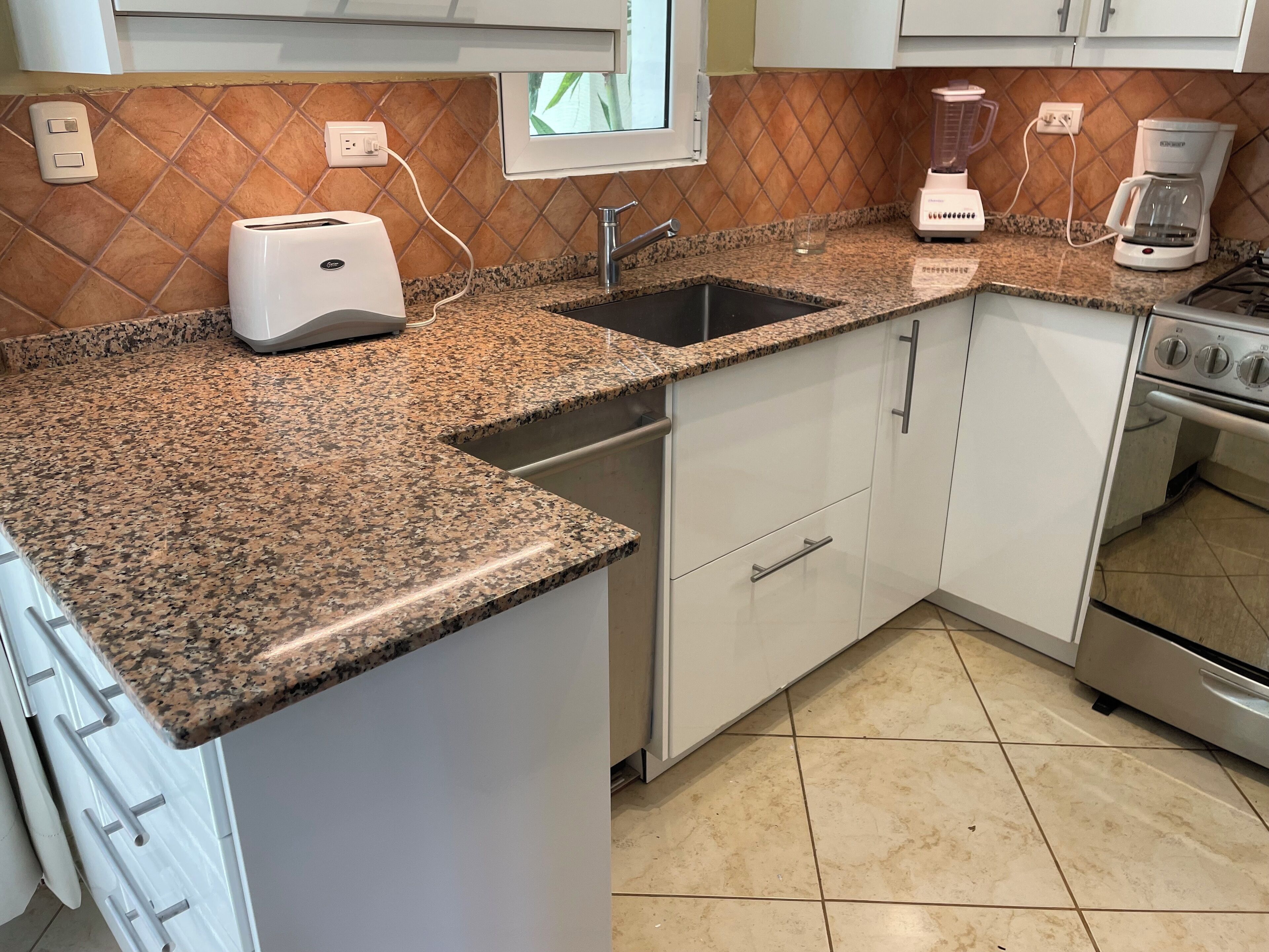 Una cocina con encimeras de granito, gabinetes blancos, un lavamanos de acero inoxidable, un tostador, una licuadora y una cafetera.