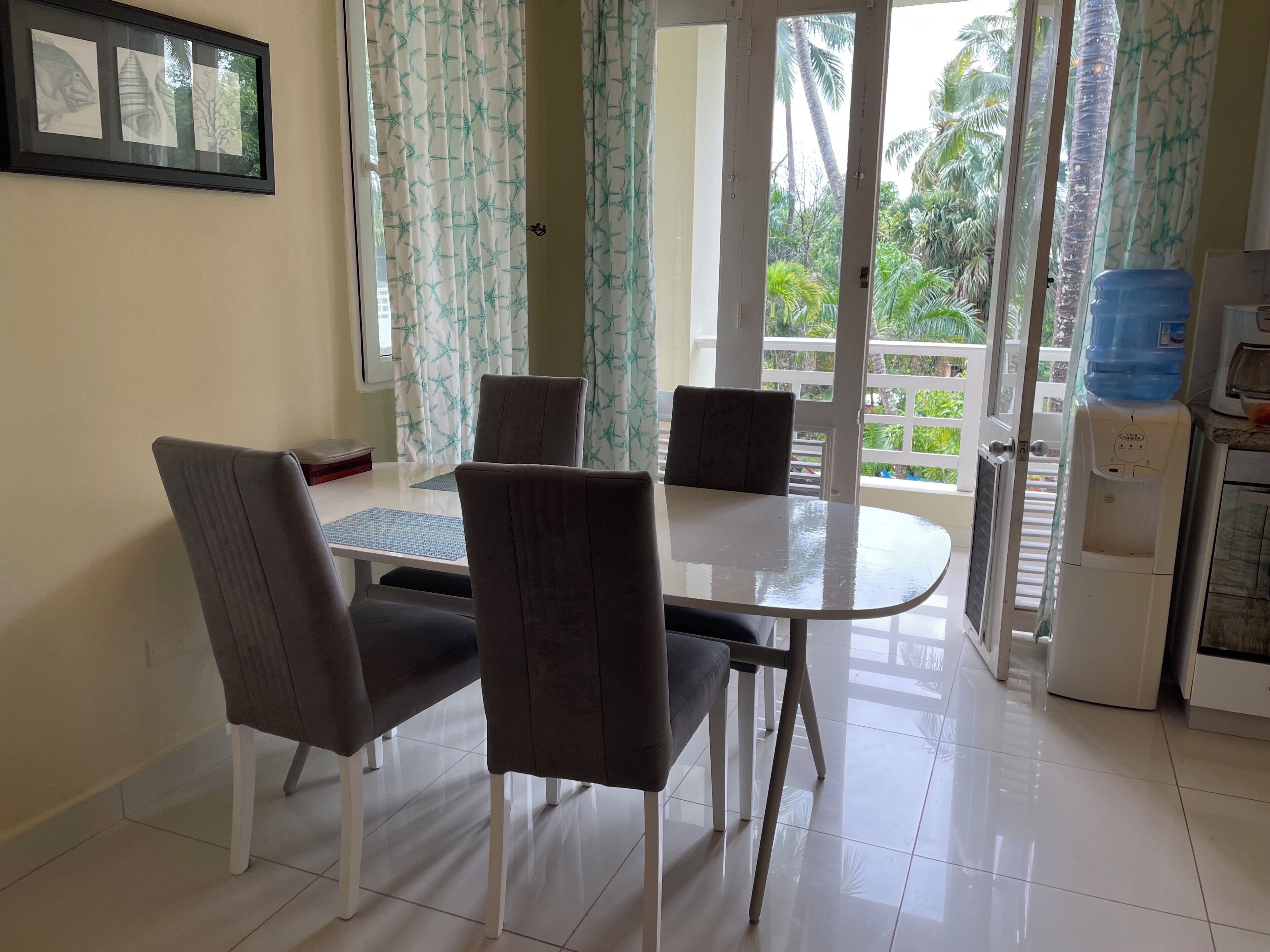 Un comedor con una mesa, sillas, un dispensador de agua y vistas al exterior con vegetación.