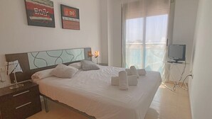 Una cama bien tendida con sábanas y toallas blancas, una lámpara de noche, una mesita con lámpara y un televisor sobre un mueble.