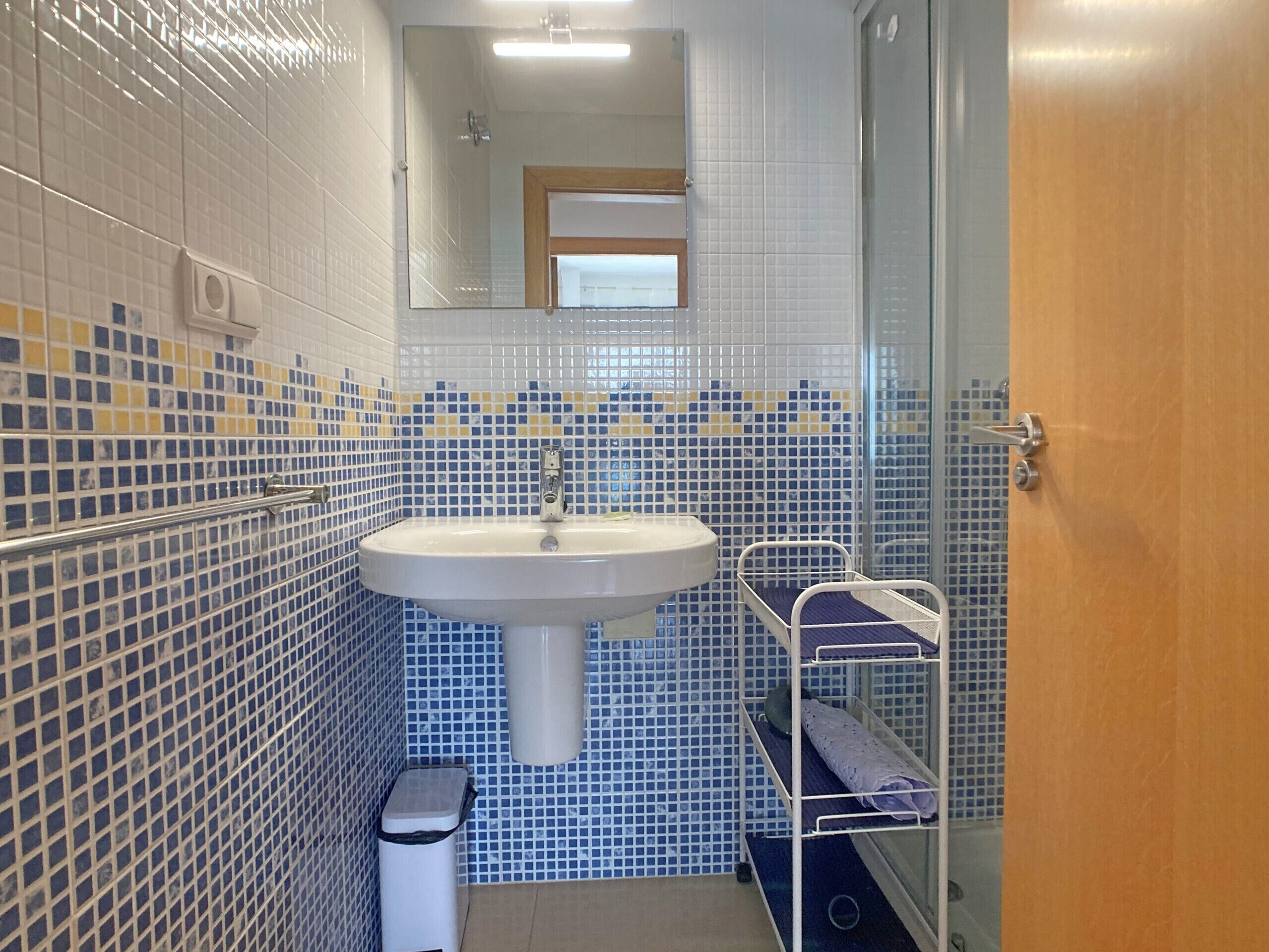 A bathroom with a white sink, tiled walls, a mirror, a glass shower enclosure, and a metal shelf unit.