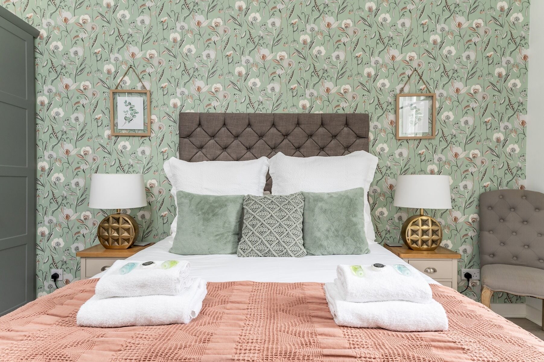 A neatly made bed with a pink quilt, two white towels, and a green and white patterned wall.