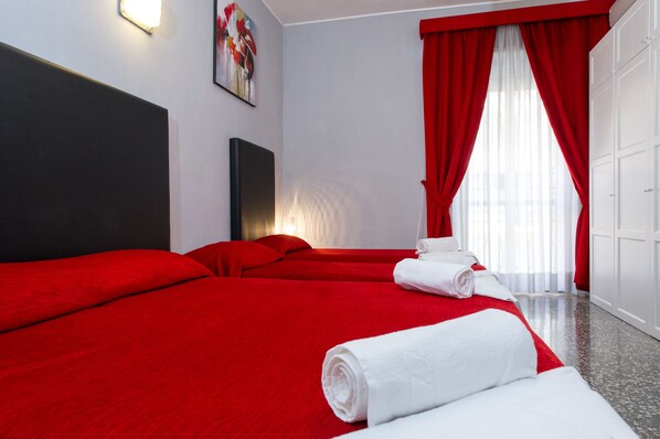 A hotel room with two beds, red bedding, white towels, a painting on the wall, and a large window with red curtains.