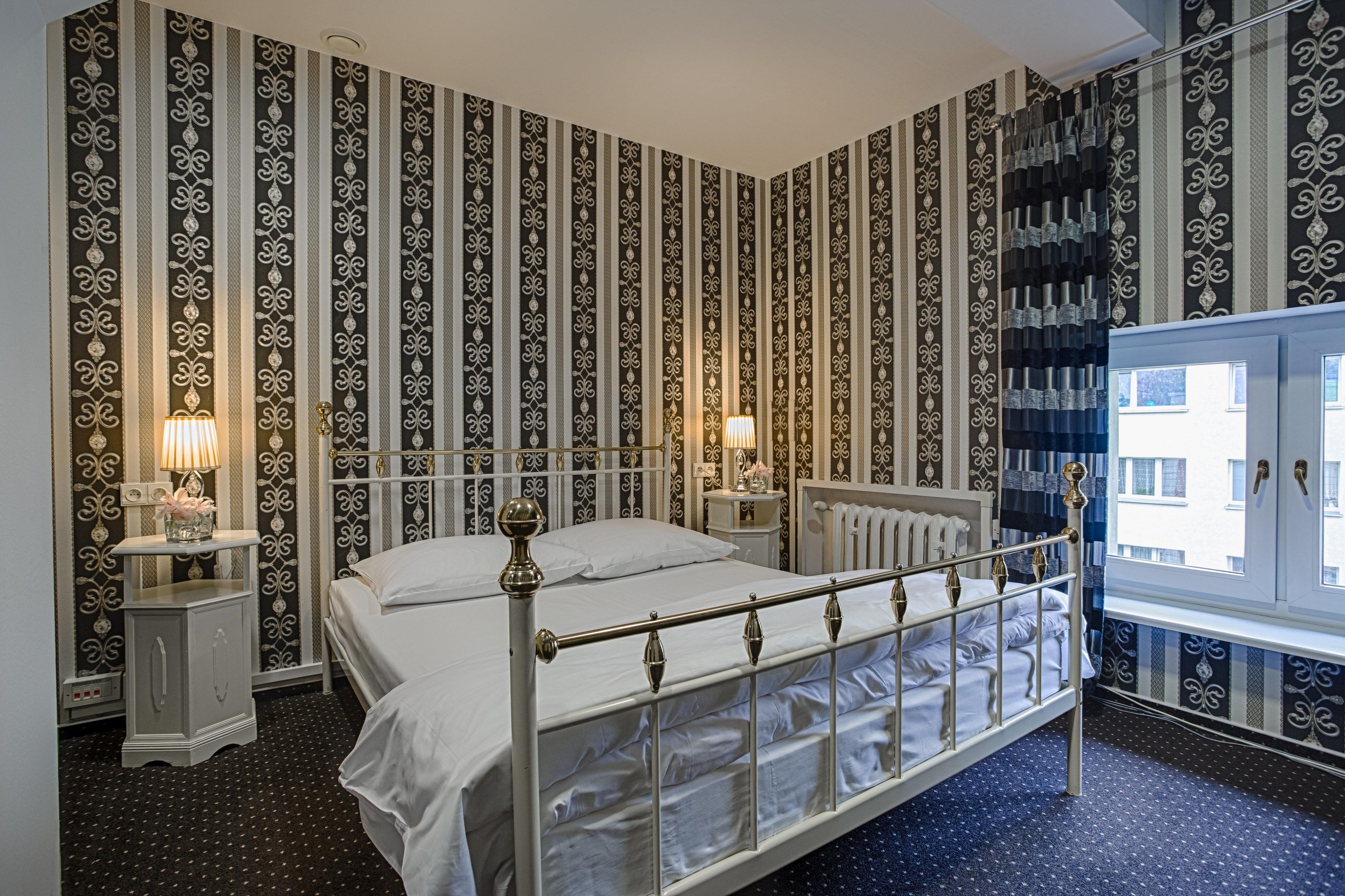 A bedroom with a metal bed frame, two bedside tables with lamps, patterned wallpaper, and a window with a view of buildings.