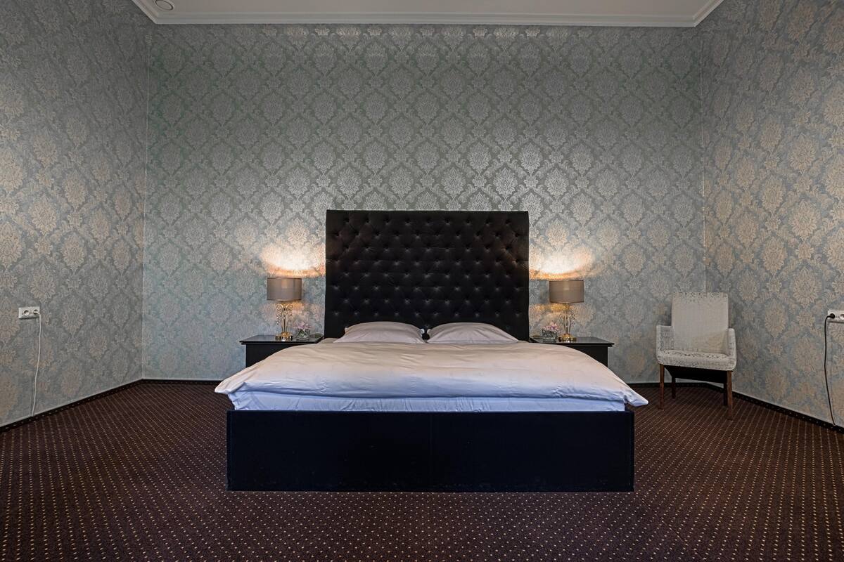 A neatly made bed with a dark headboard, two bedside lamps, a chair, and patterned wallpaper.