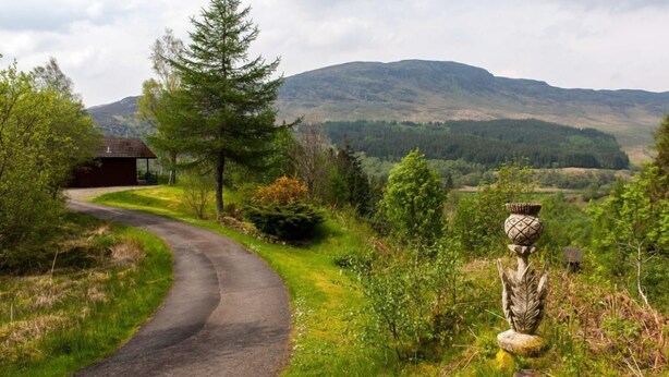 A rural road curving through a green landscape with a small building and a decorative stone structure. - Beautiful 4 star 2 bedroom chalet with stunning loch and mountain views (Crianlarich)
