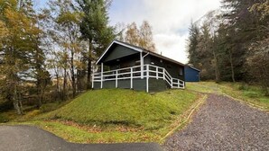 A cabin with a porch and a small deck, surrounded by trees and a grassy hill. - Beautiful 4 star 2 bedroom chalet with stunning loch and mountain views (Crianlarich)
