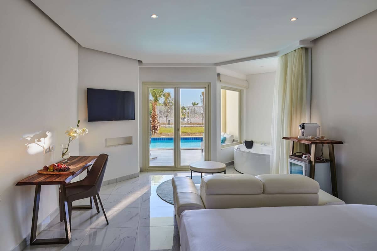A hotel room with a bed, a desk with a chair, a TV mounted on the wall, a small table with a lamp, and a view of a pool through a glass door.