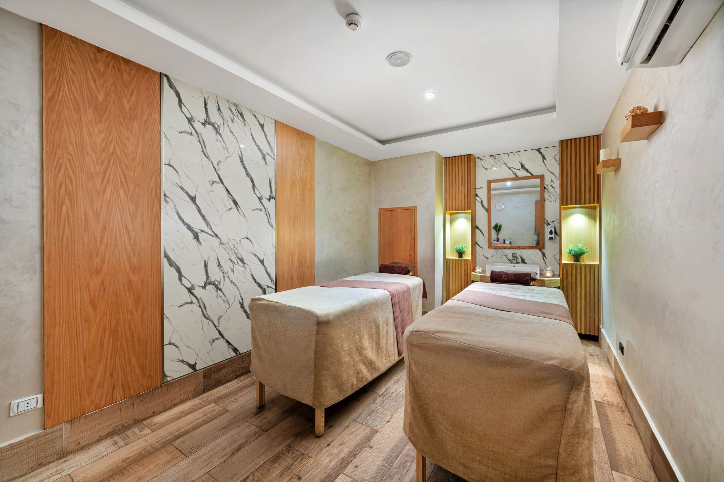 A hotel room with two beds, a wooden floor, marble wall, and a mirror.