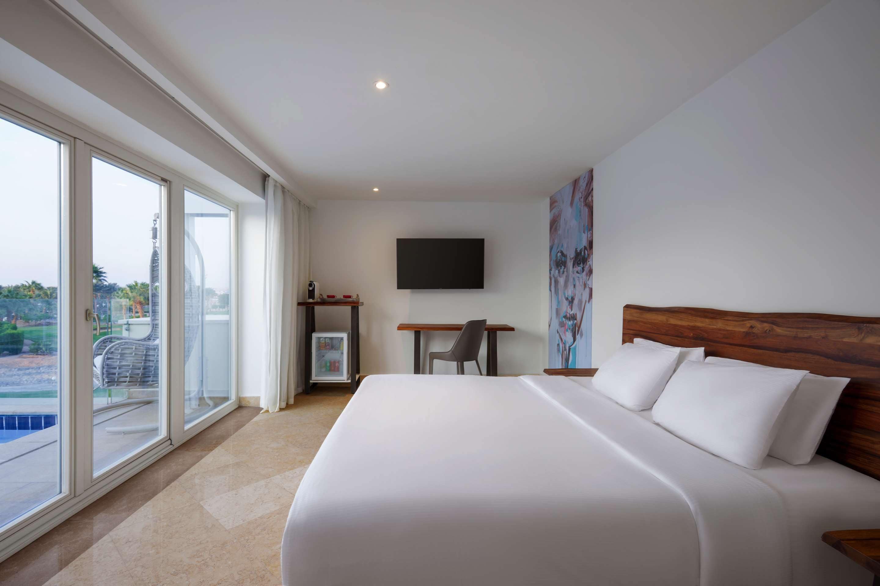 A hotel room with a large bed, a desk, a chair, a TV, and a balcony with a pool view.