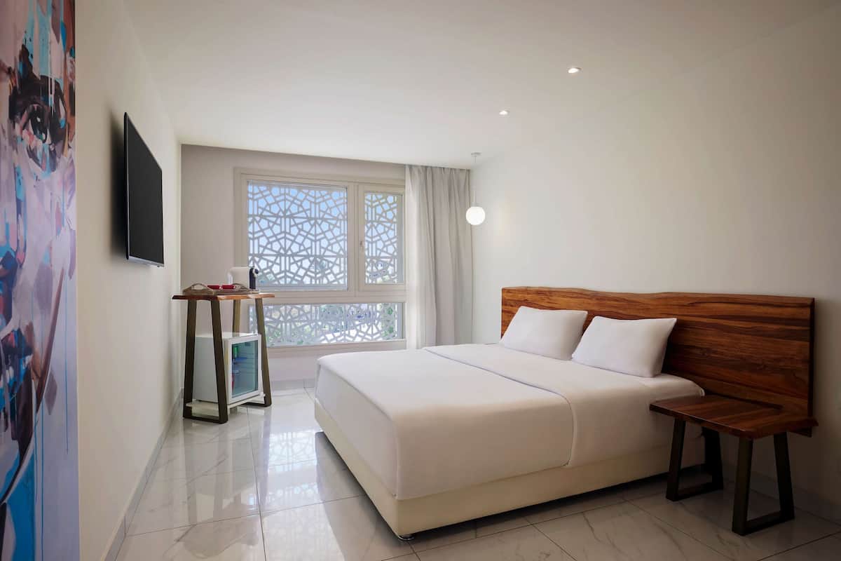 A hotel room with a large bed, a bedside table, a TV mounted on the wall, and a window with a decorative screen.
