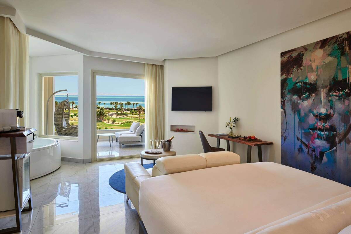 A hotel room with a large bed, a desk, a chair, a TV, and a balcony with a view of the sea.