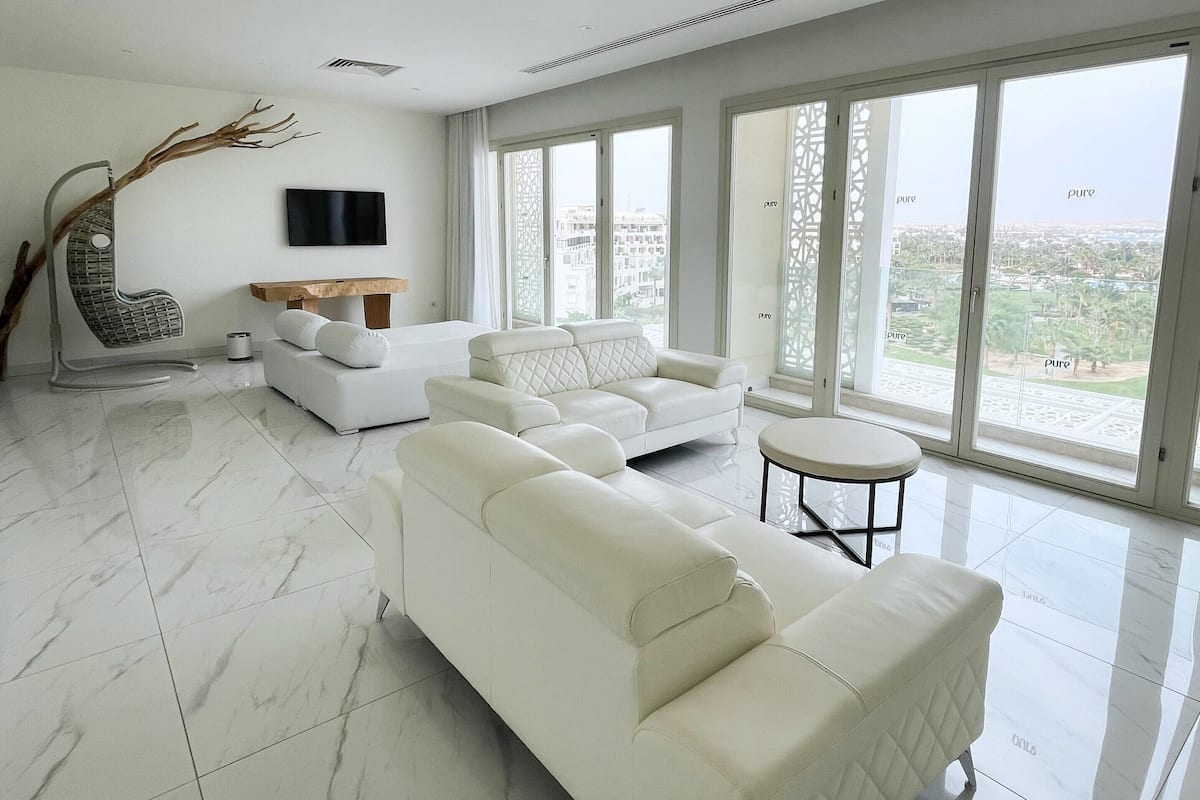 A spacious living room with a large white sofa, a small round table, and a flat-screen TV mounted on the wall.