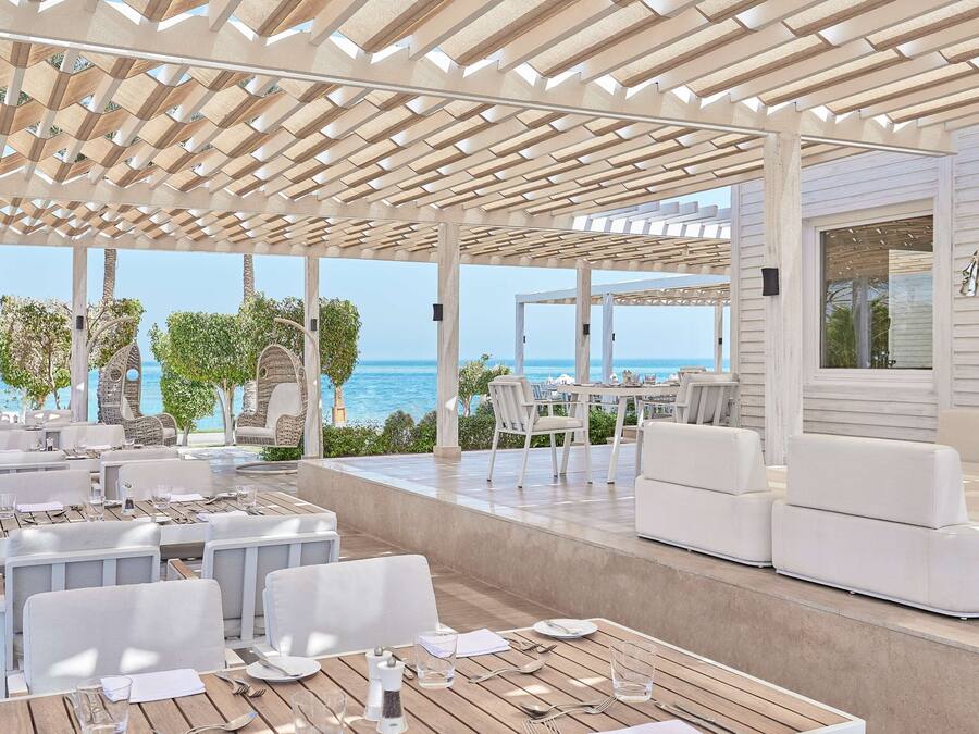 An outdoor dining area with wooden tables and white chairs, overlooking the sea.