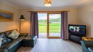 A living room with a leather sofa, a television, and a sliding glass door leading to a garden. - 4 star, 4 bedroom, 3 bathroom Scottish cottage with sensational loch and mountain views (Crianlarich)