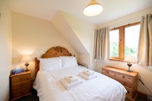 A bedroom with a wooden headboard, a bed with white linens, two bedside tables with lamps, and a window with curtains.