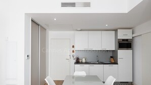 A modern kitchen with white cabinets, a built-in oven, and a dining area with a glass table and white chairs. - Sparkle-2189-Contemporary one-bedroom flat in the city centre (Cannes)