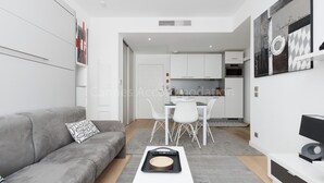 A modern living room with a grey sofa, a white coffee table, and a dining area with a white table and chairs. There is a kitchen visible in the background with white cabinets and a black countertop. - Sparkle-2189-Contemporary one-bedroom flat in the city centre (Cannes)