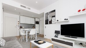 A modern living room with a grey sofa, a white coffee table, and a flat-screen TV. There is a dining area with a white table and chairs, and a kitchen in the background. - Sparkle-2189-Contemporary one-bedroom flat in the city centre (Cannes)