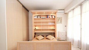 Un dormitorio con estantería de madera empotrada, una cama con dos almohadas, un radiador y una ventana con cortinas.