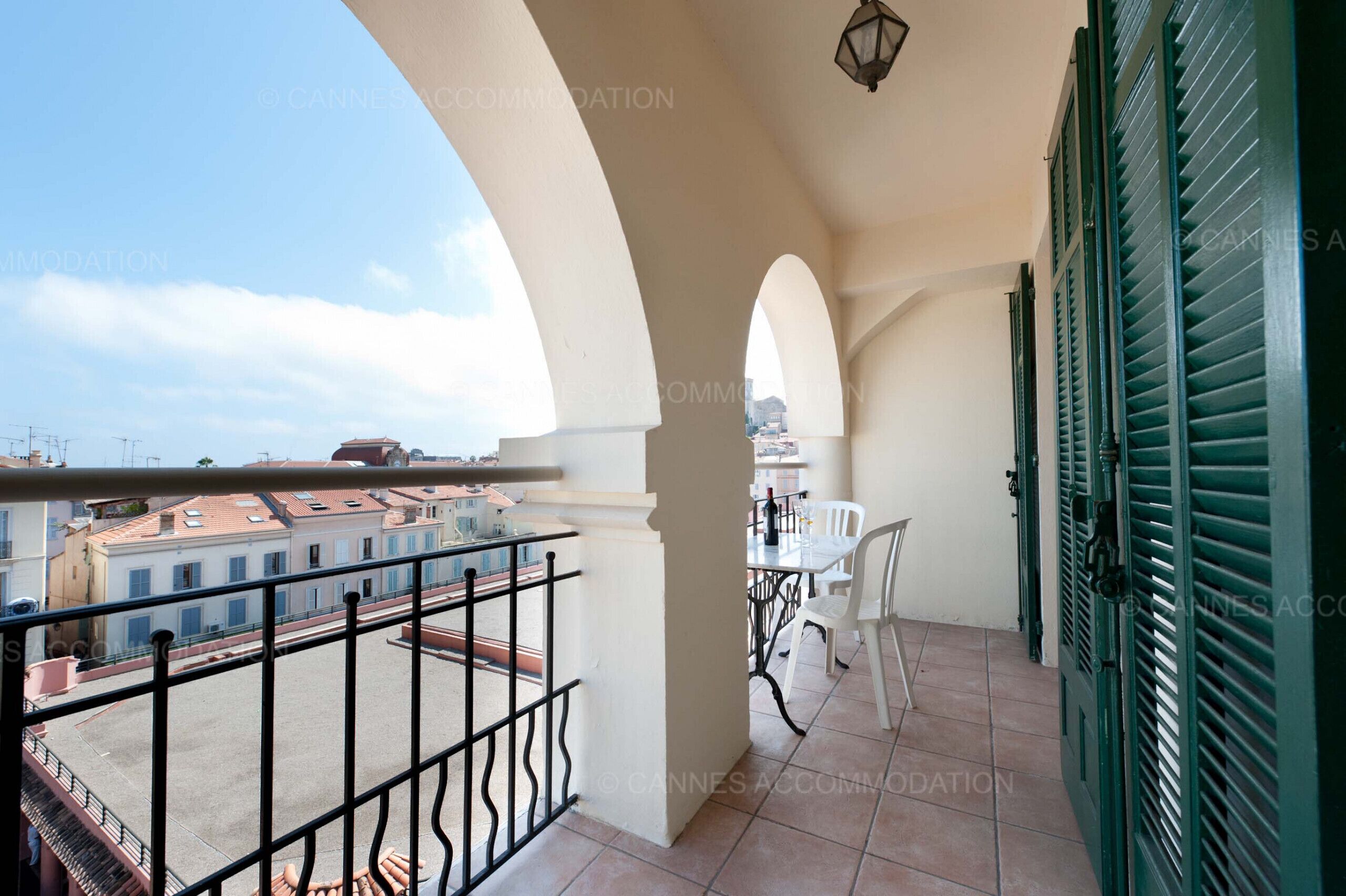 Un balcon donnant sur des immeubles, une table avec des chaises et des volets verts.