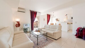 Ein modernes Wohnzimmer mit Sofa, Ohrensessel und Couchtisch. Im Hintergrund ist eine Küche mit Mikrowelle und Schränken zu sehen.